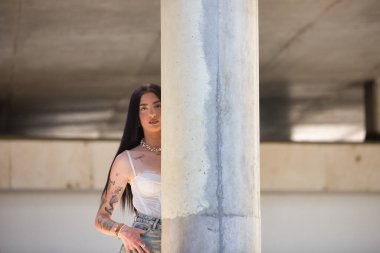 Kollarında dövmeleri olan ve sıradan kıyafetler giymiş genç, çekici bir Latin kadın beton bir sütunun arkasına saklanıyor. Kadın makyaj yapmış.