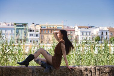 Kollarında ve bacaklarında dövmeleri olan, kahverengi elbisesi ve siyah çizmeleri olan genç ve çekici bir Latin kadın kafasına dokunuyor. Kadın taş bir duvarda oturuyor. Arka planda nehir ve evler