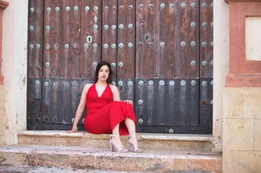 Uzun siyah saçlı, kırmızı giyinmiş, yüksek topuklu giyen, kilisenin merdivenlerinde oturan ve tahta kapıya dayanan güzel bir genç kız.