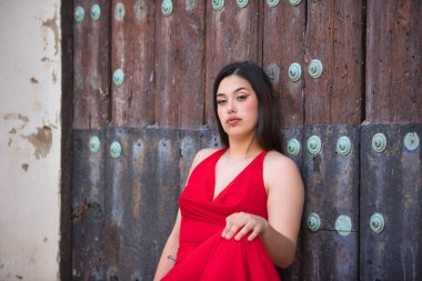 Uzun siyah saçlı, kırmızı giyinmiş, yüksek topuklu giyen, kilisenin merdivenlerinde oturan ve tahta kapıya dayanan güzel bir genç kız.