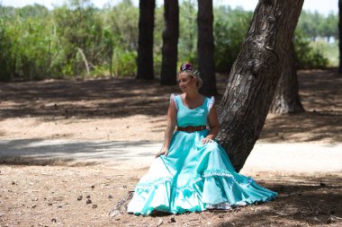 Geleneksel yeşil flamenko elbisesi giymiş, saçında bir çiçekle genç ve güzel bir İspanyol kadın tipik bir Endülüs haccında bir çam ağacının gövdesinde duruyor.