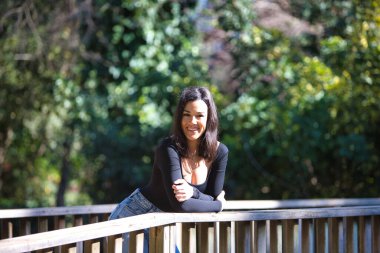 Esmer bir kadın, genç ve güzel günlük giysiler içinde tahta bir köprünün parmaklıklarına yaslanmış. Kadın farklı vücut duruşları sergiliyor. Arka planda yeşil bitki örtüsü