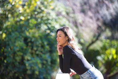 Esmer bir kadın, genç ve güzel günlük giysiler içinde tahta bir köprünün parmaklıklarına yaslanmış. Kadın farklı vücut duruşları sergiliyor. Arka planda yeşil bitki örtüsü