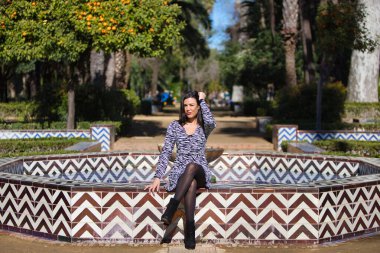 Altın sekizgen seramik fıskiyenin kenarında oturan zebra desenli hayvan desenli elbiseli güzel esmer kadın. Kadın bacak bacak üstüne atmış ve güneşli bir günde parkta mutlu.