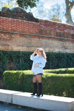 Sarışın, genç ve güzel bir kadın. Taş bir bankta günlük giysiler içinde. Kadın manken gibi farklı vücut duruşları sergiliyor ve kameraya bakıyor.