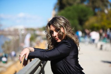 Siyah giyinmiş genç, güzel sarışın kadın gri metal korkuluklara yaslanıyor. Kadın mutlu ve kameraya bakarak gülümsüyor.