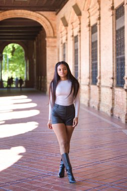 Deri şort, siyah çizme ve bej bluz giyen genç ve güzel Afrikalı kadın tatilde ve İspanya 'nın ünlü meydanında Seville' de yürüyor. Seyahat ve tatil konsepti