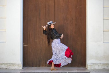 Latin bir kadın, Endülüs 'ün tipik bir sokağındaki ahşap bir kapının önünde dans eden genç ve güzel bir flamenko. Kadın, siyah benekli, şapkalı ve ceketli beyaz bir elbise giyiyor.