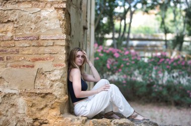 Genç, güzel, beyaz pantolon ve siyah pamuklu gömlek giyen sarışın bir kadın ormandaki yıkık dökük bir binanın taş duvarında oturuyor. Kadın dinleniyor ve eliyle kafasına dokunuyor.