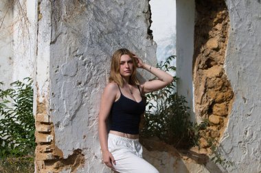 Beyaz pantolon ve siyah pamuklu gömlek giymiş genç, güzel bir sarışın kadın ormanda terk edilmiş, harap olmuş bir evin duvarına yaslanıyor. Kadın ciddi ve üzgün görünüyor..