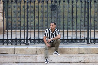 Genç, yakışıklı, siyah saçlı, çizgili gömlek ve kargo pantolonu giyen bir adam Seville, Endülüs 'teki Gotik katedralin önünde oturuyor. Adam şehirde tatilde.