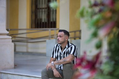 Çizgili gömlek ve kargo pantolonu giyen sakallı genç, yakışıklı bir adam bir binanın beyaz mermer merdiveninde oturuyor. Adam etrafa bakıyor.