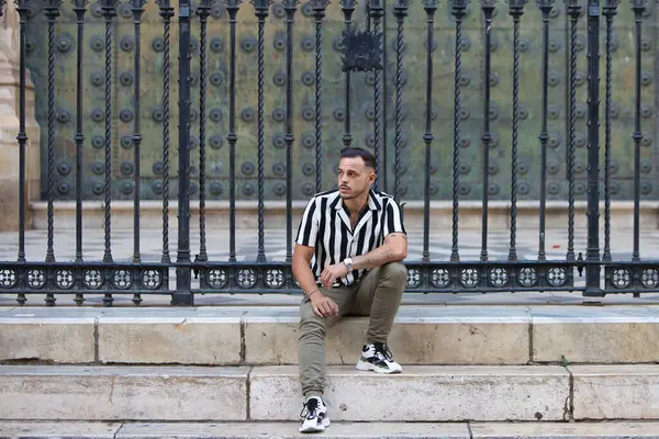 Genç, yakışıklı, siyah saçlı, çizgili gömlek ve kargo pantolonu giyen bir adam Seville, Endülüs 'teki Gotik katedralin önünde oturuyor. Adam şehirde tatilde.