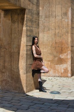 Latin bir kadın, genç ve güzel esmer, kollarında ve bacaklarında dövmeleri olan, kahverengi bir elbise ve siyah çizmeler giyen, kahverengi beton bir duvara yaslanan. Kadın rahatladı.