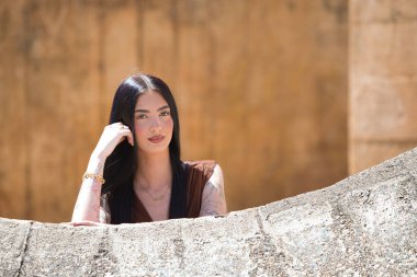 Uzun saçlı, genç, güzel esmer bir kadın yanağını elinde dinlendiriyor ve düşünceli ve bitkin görünüyor. Kadın farklı yönlere bakıyor. Arka planda kahverengi bir duvar var