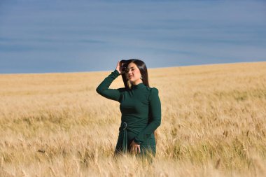 Yeşil giyinmiş, güzel, koyu renk saçlı Latin bir kız yaz günü kuru, sarı bir buğday tarlasının arasında duruyor. Arka planda, mavi gökyüzü