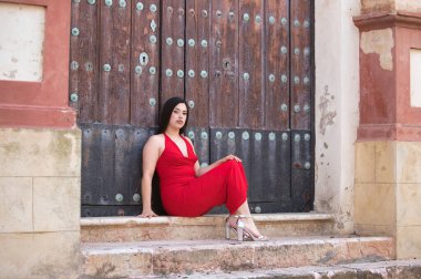 Uzun siyah saçlı, kırmızı giyinmiş, yüksek topuklu giyen, kilisenin merdivenlerinde oturan ve tahta kapıya dayanan güzel bir genç kız.