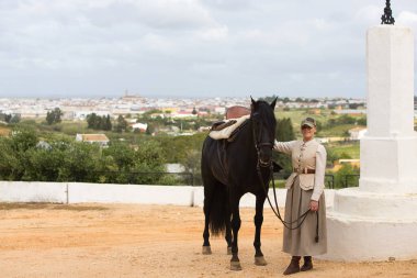 Genç ve güzel bir kadın siyah safkan İspanyol atının yüzünü sevgiyle tutuyor ve kameraya mutlulukla bakıyor. Kadın tipik bir atlı kadın kostümü giymiş.