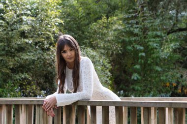 Beyaz örgü elbiseli, genç ve güzel esmer kadın, parktaki bir köprünün ahşap korkuluklarına yaslanarak farklı ifadeler sergiliyor. Arka planda ağaçlar ve bitki örtüsü