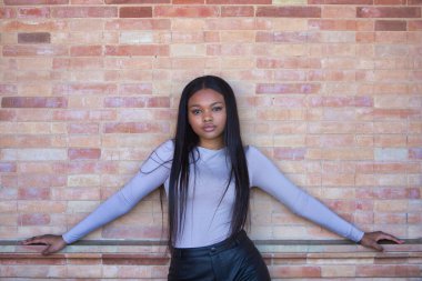 Afrikalı bir kadın, genç ve güzel, bej bluzlu ve siyah deri şortlu. Kolları açık, kırmızı tuğladan bir duvara yaslanmış.