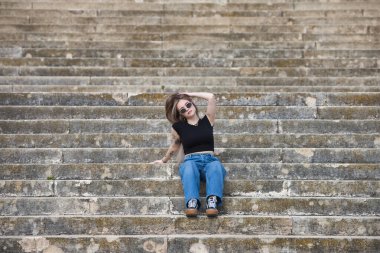 Genç ve güzel sarışın kadın, dövmeli ve kot pantolon, siyah gömlek ve güneş gözlüğü takıyor, merdivenlerde oturuyor ve farklı duruşlar ve ifadeler sergiliyor.