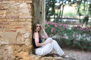 Genç, güzel, beyaz pantolon ve siyah pamuklu gömlek giyen sarışın bir kadın ormandaki yıkık dökük bir binanın taş duvarında oturuyor. Kadın dinleniyor ve eliyle kafasına dokunuyor.