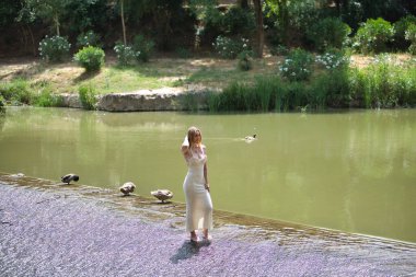 Bej örülü bir elbise giyen genç, güzel bir sarışın kadın ördekler ve bitkilerle dolu bir nehirde küçük bir çağlayan oluşturan bir şelalede yalın ayak yürüyor.