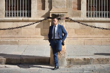 Genç, yakışıklı, koyu renk saçlı, takım elbiseli, kravatlı ve siyah ayakkabılı bir işadamı elleri cebinde, Seville 'in eski kasabasında zincirli bir sütuna yaslanmış sabırsız bir şekilde bekliyor.