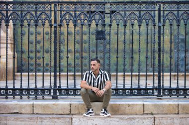 Genç, yakışıklı, siyah saçlı, çizgili gömlek ve kargo pantolonu giyen bir adam Seville, Endülüs 'teki Gotik katedralin önünde oturuyor. Adam şehirde tatilde.