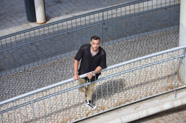 Sakallı, siyah tişörtlü ve bej kargo pantolonlu genç, yakışıklı bir adam, metal bir korkuluğa tutunuyor, rampadan aşağı indi ve yukarı bakıyor. Fotoğraf yukarıdan çekildi