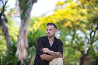 Sakallı, dövmeli, küpeli ve modern elbiseli genç, yakışıklı bir eşcinsel parkta, kollarını bacağının üzerine koyuyor, kollarını bankın üzerine dayamış, ve ciddi bir şekilde yan tarafa bakıyor.