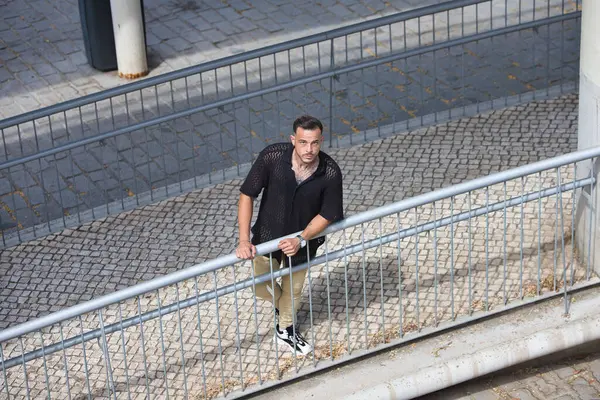 Sakallı, siyah tişörtlü ve bej kargo pantolonlu genç, yakışıklı bir adam, metal bir korkuluğa tutunuyor, rampadan aşağı indi ve yukarı bakıyor. Fotoğraf yukarıdan çekildi
