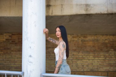 Latin kadın, genç ve güzel esmer, kollarında dövmeleri var ve beton bir sütuna yaslanmış günlük kıyafetler giyiyor. Kadın makyaj yapmış.