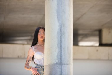 Kollarında dövmeleri olan ve sıradan kıyafetler giymiş genç, çekici bir Latin kadın beton bir sütunun arkasına saklanıyor. Kadın makyaj yapmış.