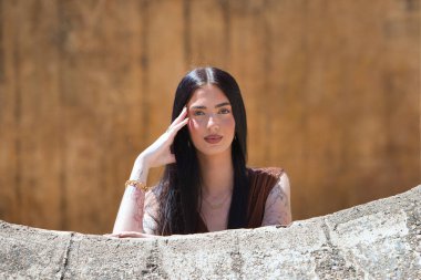 Uzun saçlı, genç, güzel esmer bir kadın yanağını elinde dinlendiriyor ve düşünceli ve bitkin görünüyor. Kadın farklı yönlere bakıyor. Arka planda kahverengi bir duvar var