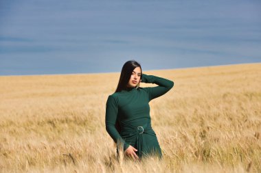 Yeşil giyinmiş, güzel, koyu renk saçlı Latin bir kız yaz günü kuru, sarı bir buğday tarlasının arasında duruyor. Arka planda, mavi gökyüzü