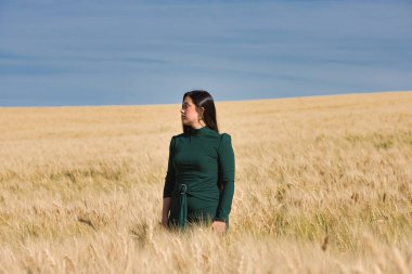 Yeşil giyinmiş, güzel, koyu renk saçlı Latin bir kız yaz günü kuru, sarı bir buğday tarlasının arasında duruyor. Arka planda, mavi gökyüzü
