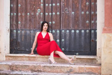 Uzun siyah saçlı, kırmızı giyinmiş, yüksek topuklu giyen, kilisenin merdivenlerinde oturan ve tahta kapıya dayanan güzel bir genç kız.