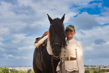 Genç ve güzel bir kadın siyah safkan İspanyol atının yüzünü sevgiyle tutuyor ve kameraya mutlulukla bakıyor. Kadın tipik bir atlı kadın kostümü giymiş.