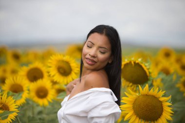 Koyu tenli, beyaz gömlekli ve çıplak omuzlu güzel bir Latin kadın güneşli bir günde ayçiçeklerinin arasında duruyor. Şehvetli kadın gülümser ve mutlu görünür.