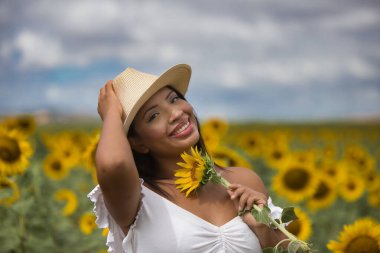 Beyaz gömlek ve hasır şapka giyip ayçiçeklerinin arasında duran koyu tenli genç bir Latin kadın çiftçinin portresi. Kız elinde bir çiçek tutuyor ve kokluyor ve ona bakıyor.