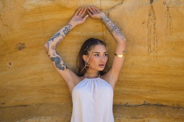 Makyaj yapan güzel bir genç kadın, bej bir elbise ve deniz kenarındaki bir uçurum duvarına dayanmış altın mücevherler. Kadın farklı pozlar ve ifadeler veriyor. Güzellik ve moda kavramı