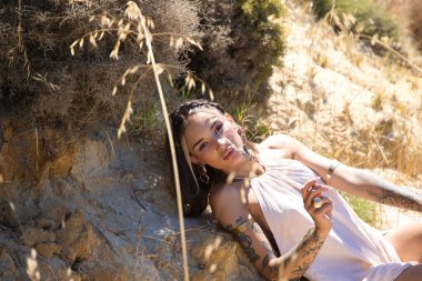 Bej bir elbise ve altın mücevherler giyen, cerrahi olarak geliştirilmiş dudakları olan genç ve güzel bir kadın, deniz kenarındaki kum tepeciklerinin arasında uzanıyor.