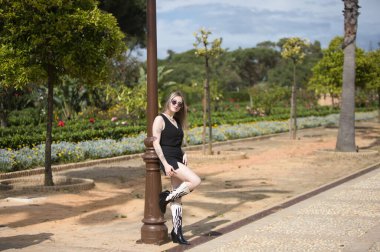 Sarışın, genç ve güzel bir kadın. Dövmeleri, güneş gözlükleri, kovboy çizmeleri ve siyah çizmeleri var. Bacağını palmiye ağaçları ve bahçeleri olan bir sokak lambasında dinlendiriyor.