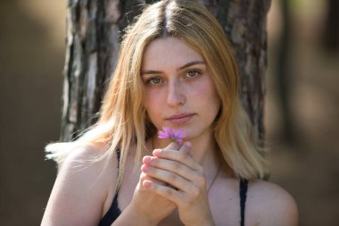 Genç, güzel, doğal sarışın bir kadın eline mor bir çiçek alıp onu kokluyor. Kadın kameraya bakıyor. Güzellik ve sağlık kavramı