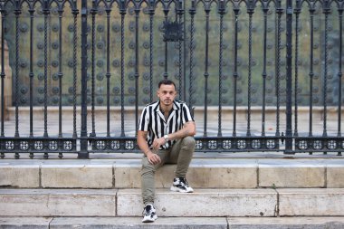 Genç, yakışıklı, siyah saçlı, çizgili gömlek ve kargo pantolonu giyen bir adam Seville, Endülüs 'teki Gotik katedralin önünde oturuyor. Adam şehirde tatilde.