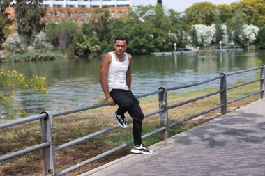 Sakallı, dövmeli, küpeli, beyaz kolsuz ve siyah pantolonlu genç, yakışıklı bir eşcinsel nehir kenarındaki metal bir korkuluğa yaslanıyor. Adam rahat ve dinleniyor.