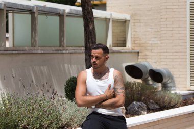 Sakallı, dövmeli, küpeli, beyaz atletli, siyah pantolonlu, çiçek tarhında oturan, kolları çaprazlanmış, ciddi görünen genç bir adam.