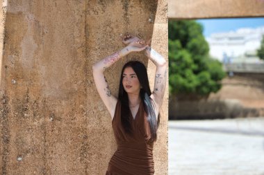 Uzun saçlı ve kahverengi elbiseli genç, çekici bir esmer kadın dövmeli kollarını kahverengi beton bir duvara doğru kaldırıyor. Kadın makyaj yapmış ve şık giyinmiş.