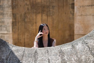 Uzun saçlı, genç, güzel esmer bir kadın yanağını elinde dinlendiriyor ve düşünceli ve bitkin görünüyor. Kadın farklı yönlere bakıyor. Arka planda kahverengi bir duvar var